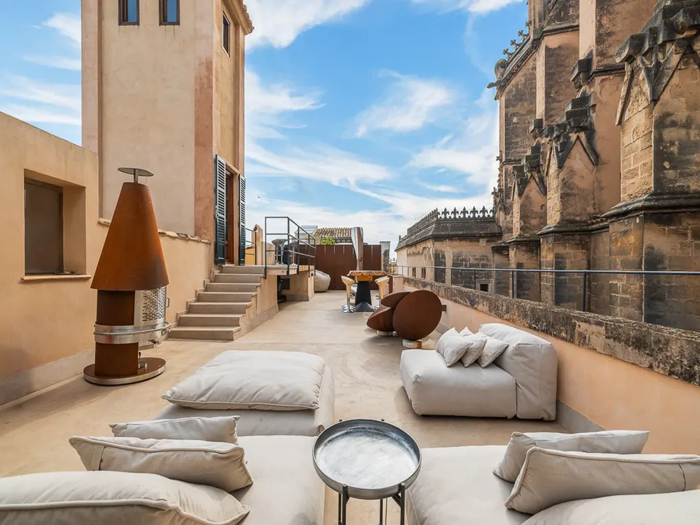 Rooftop patio with white cushions, metal table, and fireplace. Stone buildings and blue sky in the background.