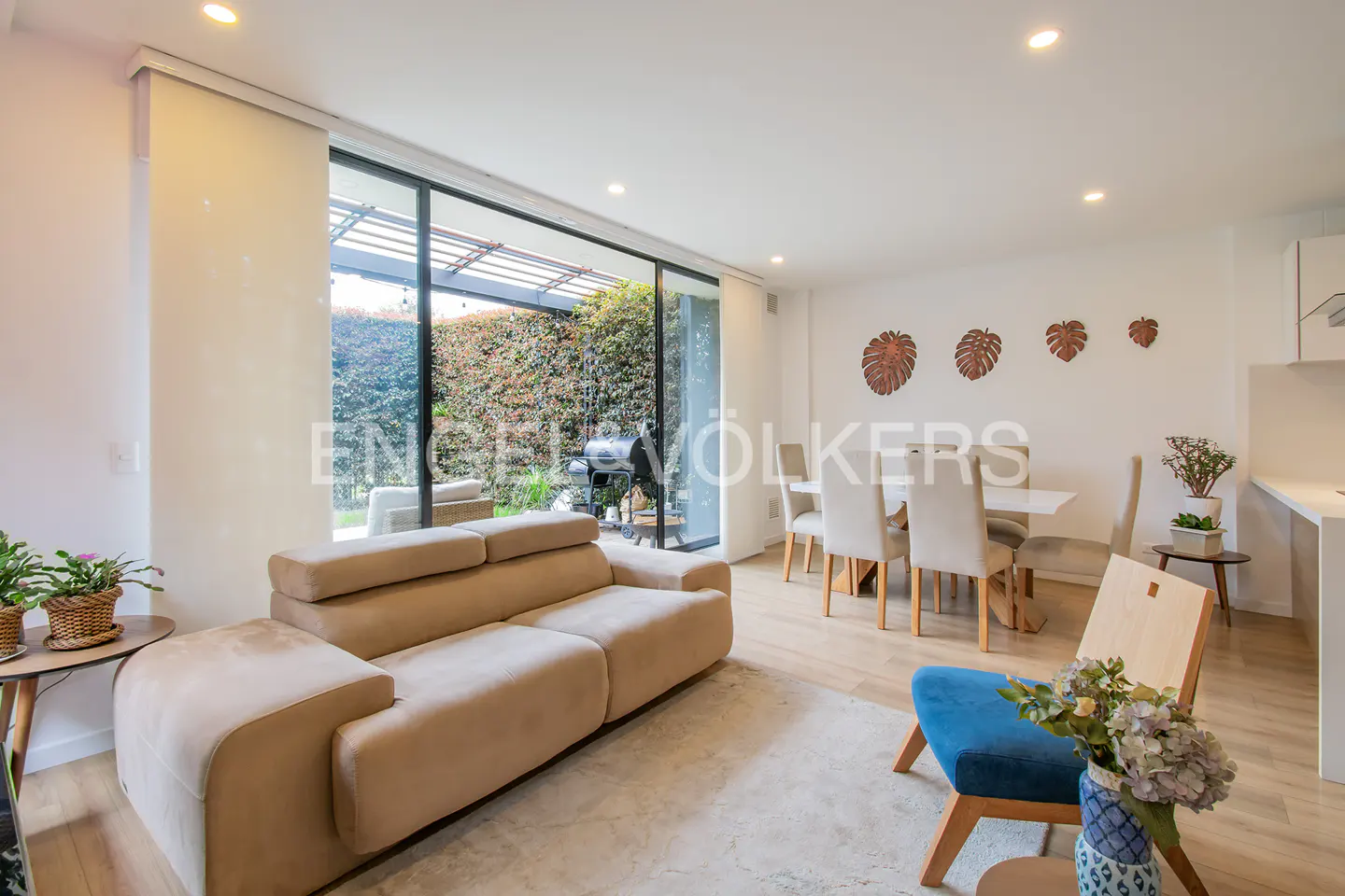 Bright living room with a beige sofa, dining table with white chairs, and a blue accent chair. A large glass door leads to a patio with greenery.