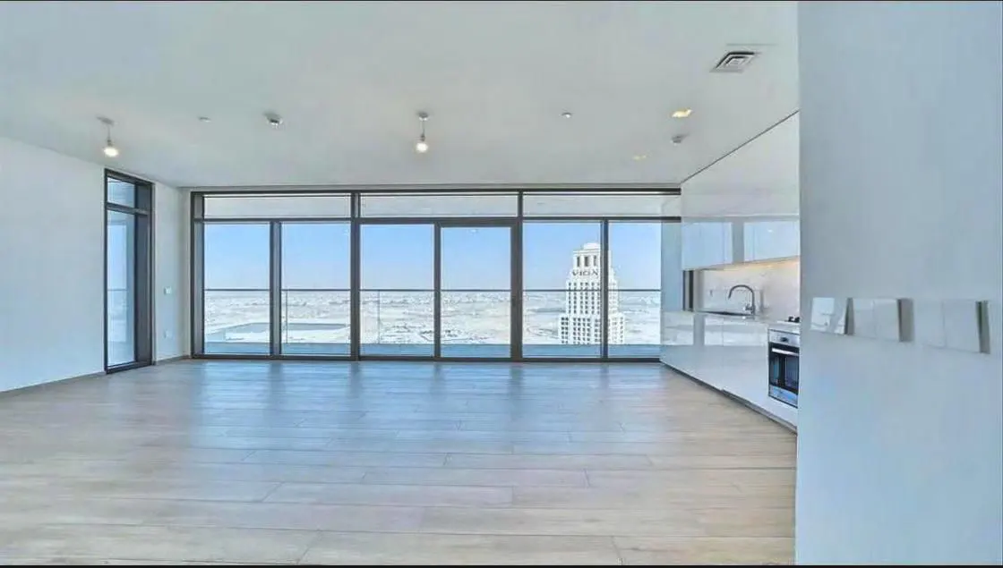 Bright, empty apartment with wood floors, white walls, and floor-to-ceiling windows. A modern white kitchen is visible. City view.