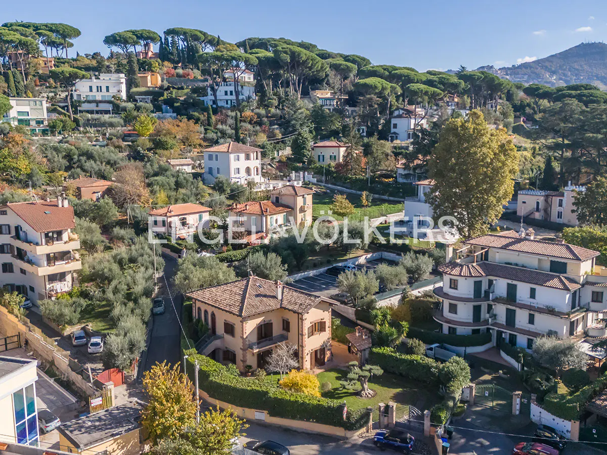 Aerial view of hillside homes with terracotta roofs, surrounded by trees and greenery under a blue sky.