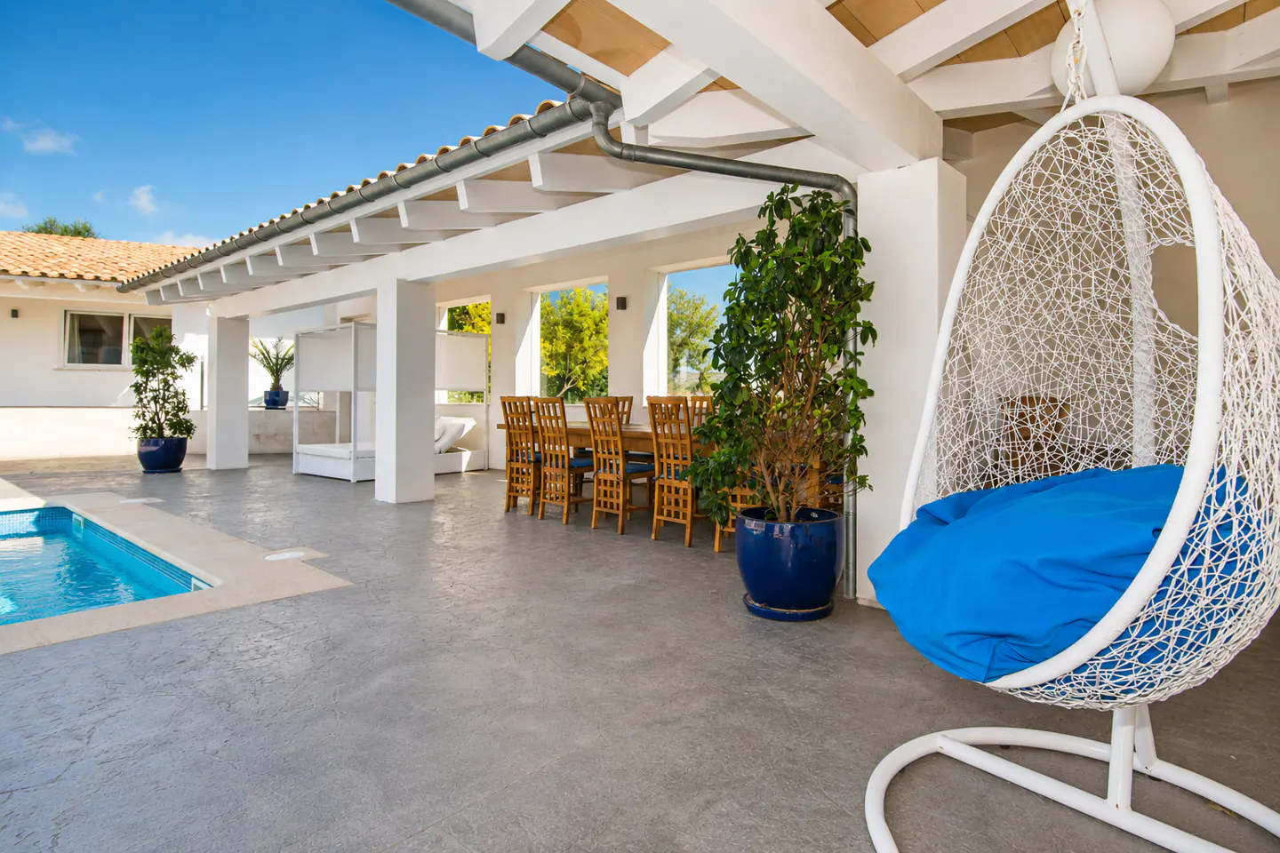 Outdoor patio with a pool, dining table, and hanging chair. The chair is white with a blue cushion. The sky is blue.
