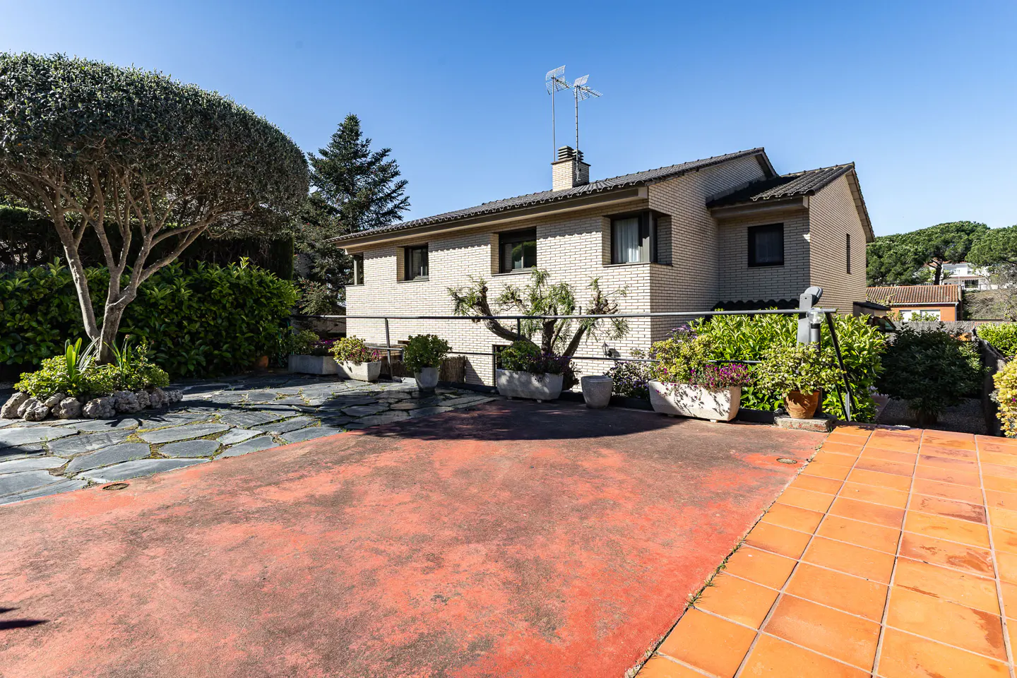Exterior view of a two-story brick house with a red patio, plants, and trees under a clear blue sky.