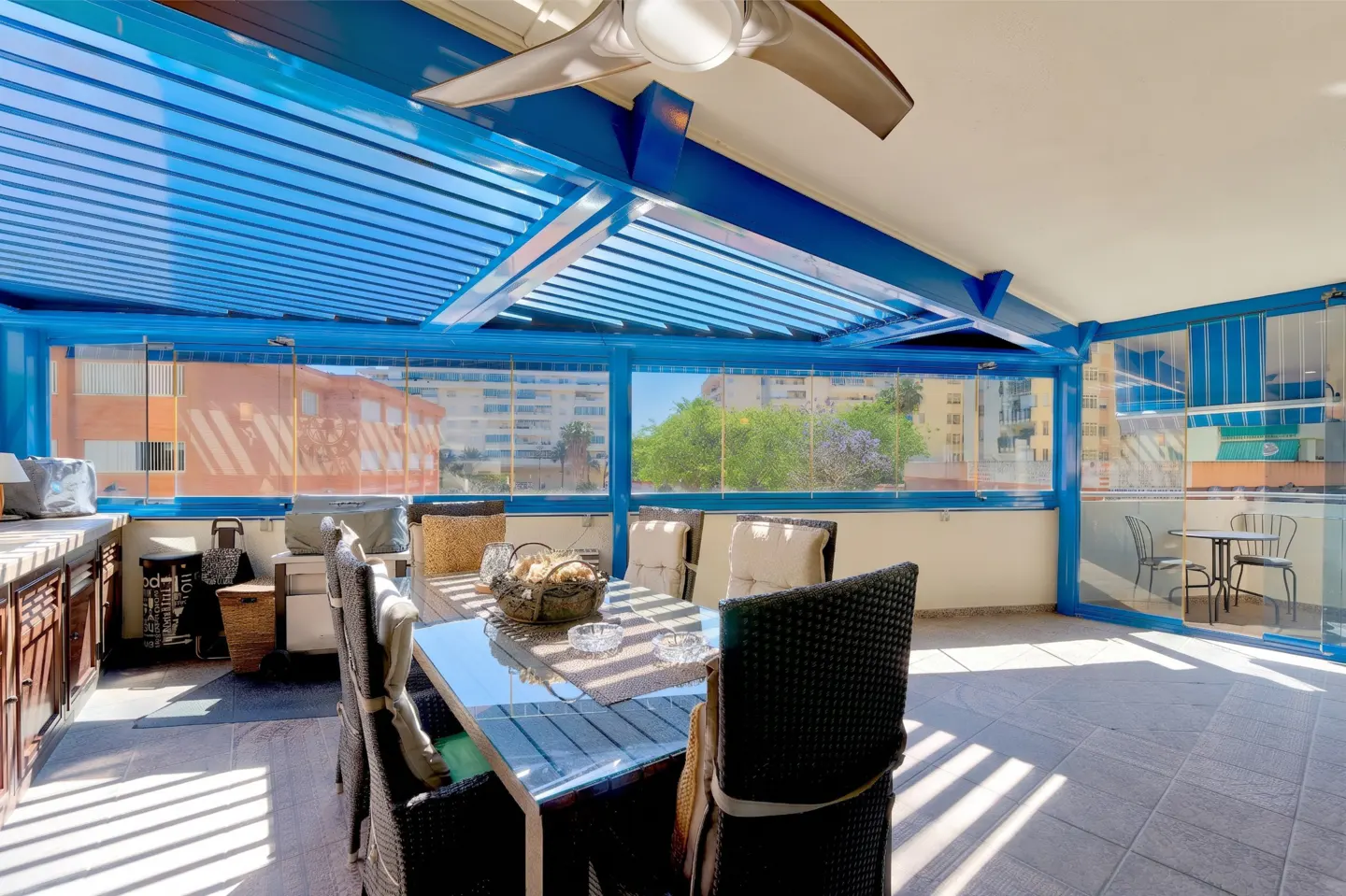 Bright sunroom with blue pergola, glass walls, and tile floor. A glass-topped table with chairs sits in the center. Outside, buildings and trees are visible.
