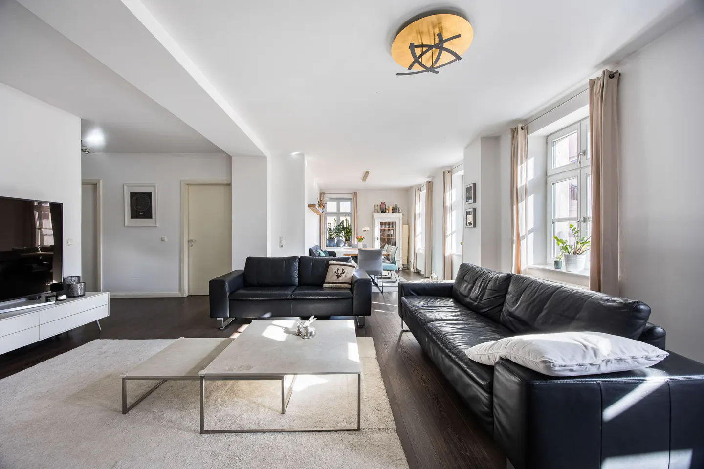 Bright living room with two black leather sofas, a white rug, and a concrete coffee table. A dining area is visible in the background.