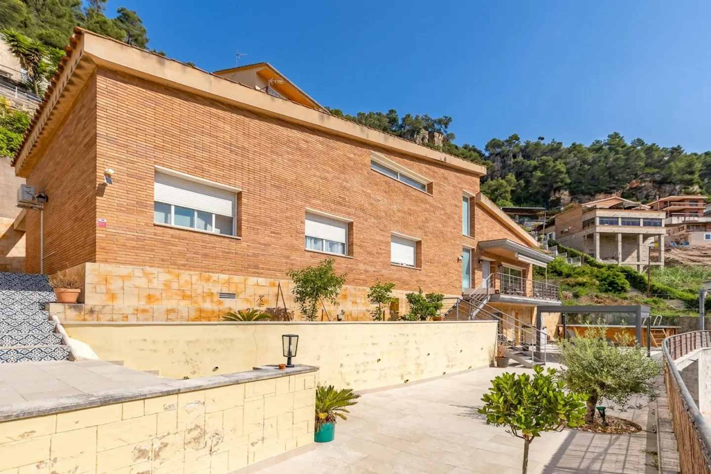 Exterior view of a two-story brick house with a patio, plants, and a view of the hillside.
