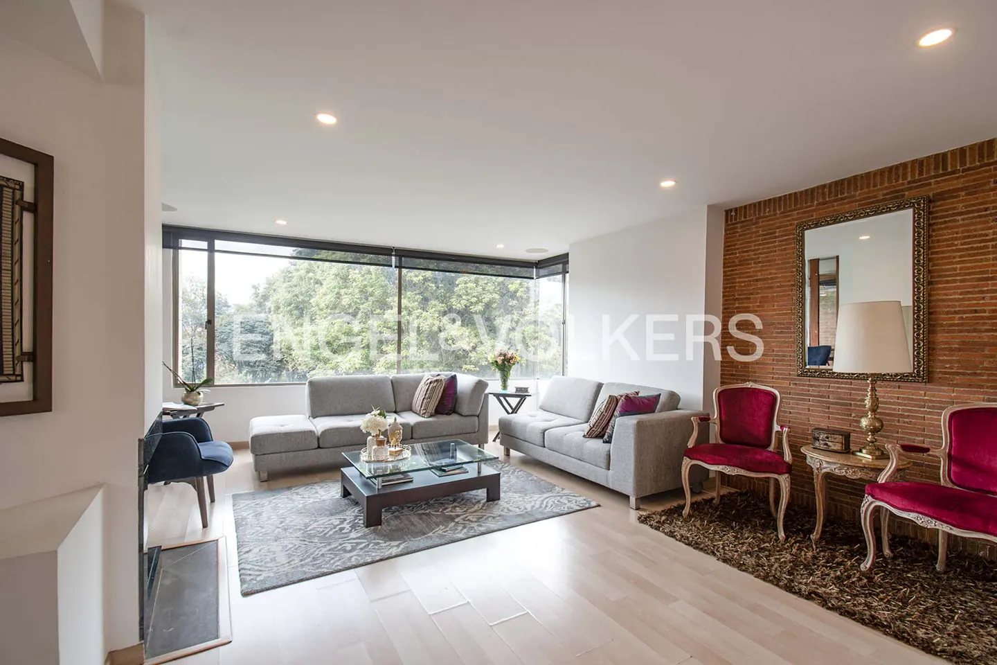 Bright living room with gray sofas, glass table, and red chairs. Large window overlooks trees. Brick wall with mirror and lamp.