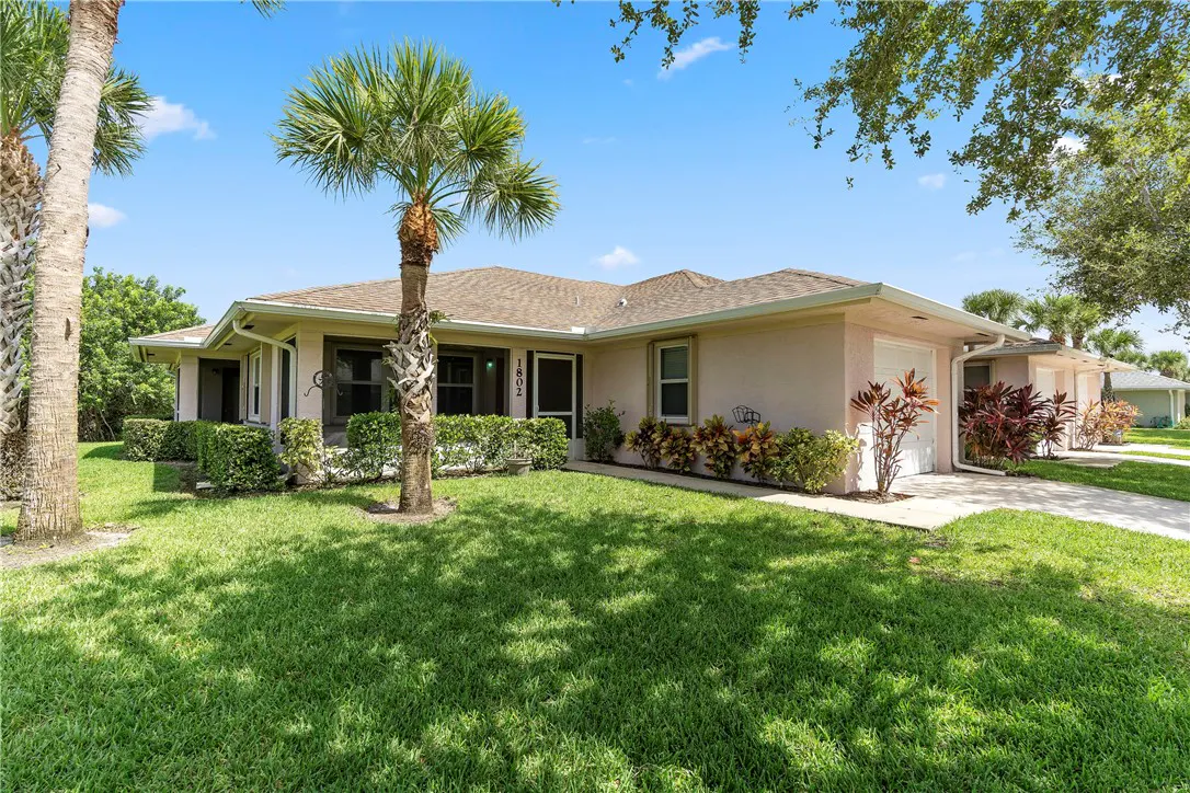 Single-story beige house with a brown roof, palm tree in the front yard, green lawn, and blue sky on a sunny day.