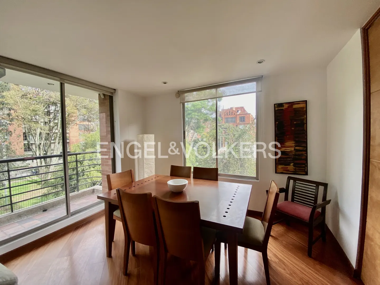 Bright dining room with wood floors, a large wood table with six chairs, and a bench. Windows overlook trees and buildings.