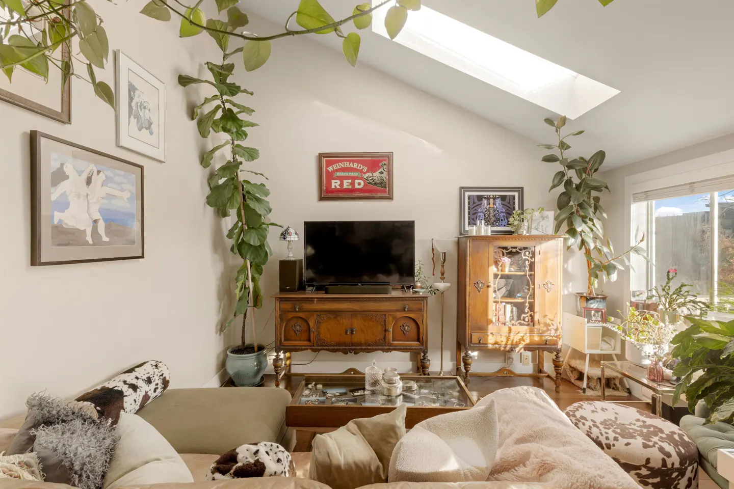 Living room with a skylight, plants, and eclectic decor. A brown wooden cabinet and TV stand are against the wall. A beige sofa is in the foreground.