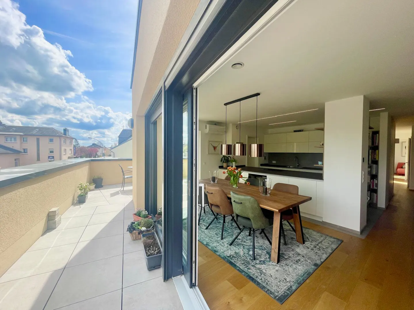 Open sliding doors reveal a modern apartment with a balcony. A dining table sits on a patterned rug, with a kitchen in the background.