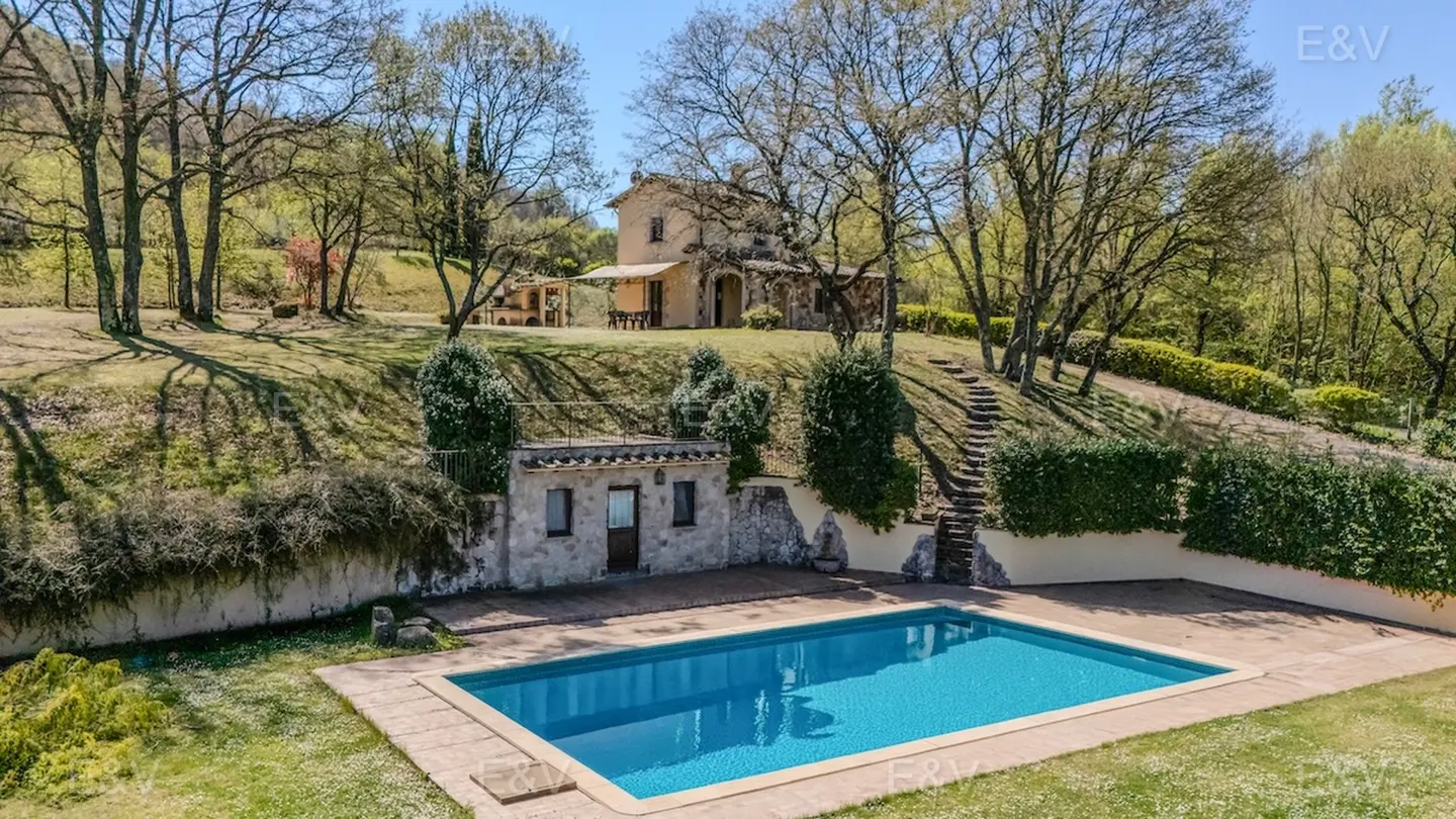 Outdoor pool with blue water, stone patio, and a small stone building. A house sits on a hill in the background, surrounded by trees.
