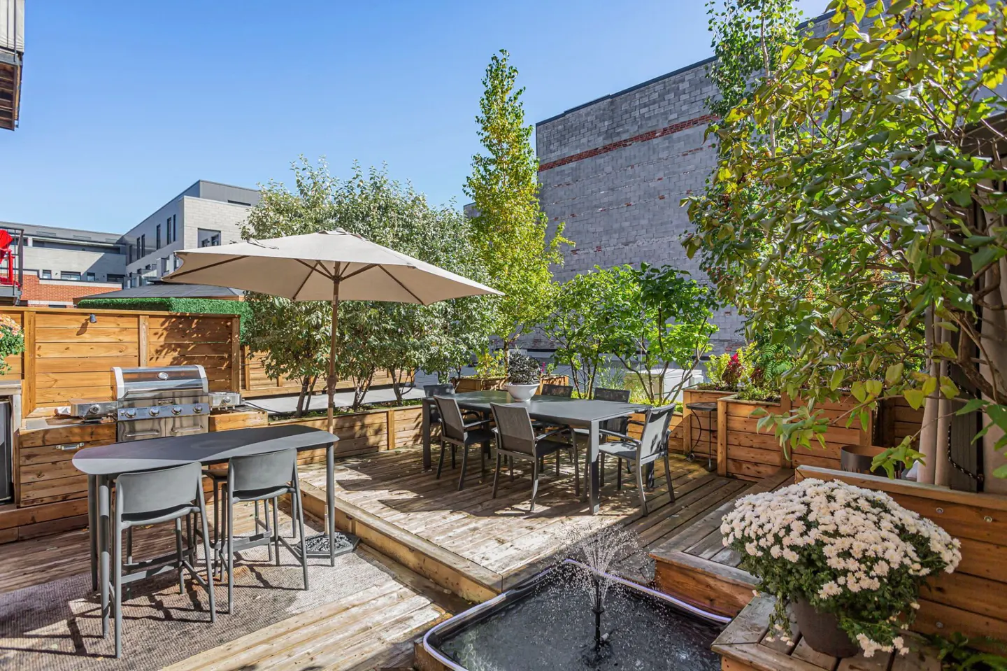 Outdoor patio with a grill, dining table with chairs, umbrella, fountain, and greenery.
