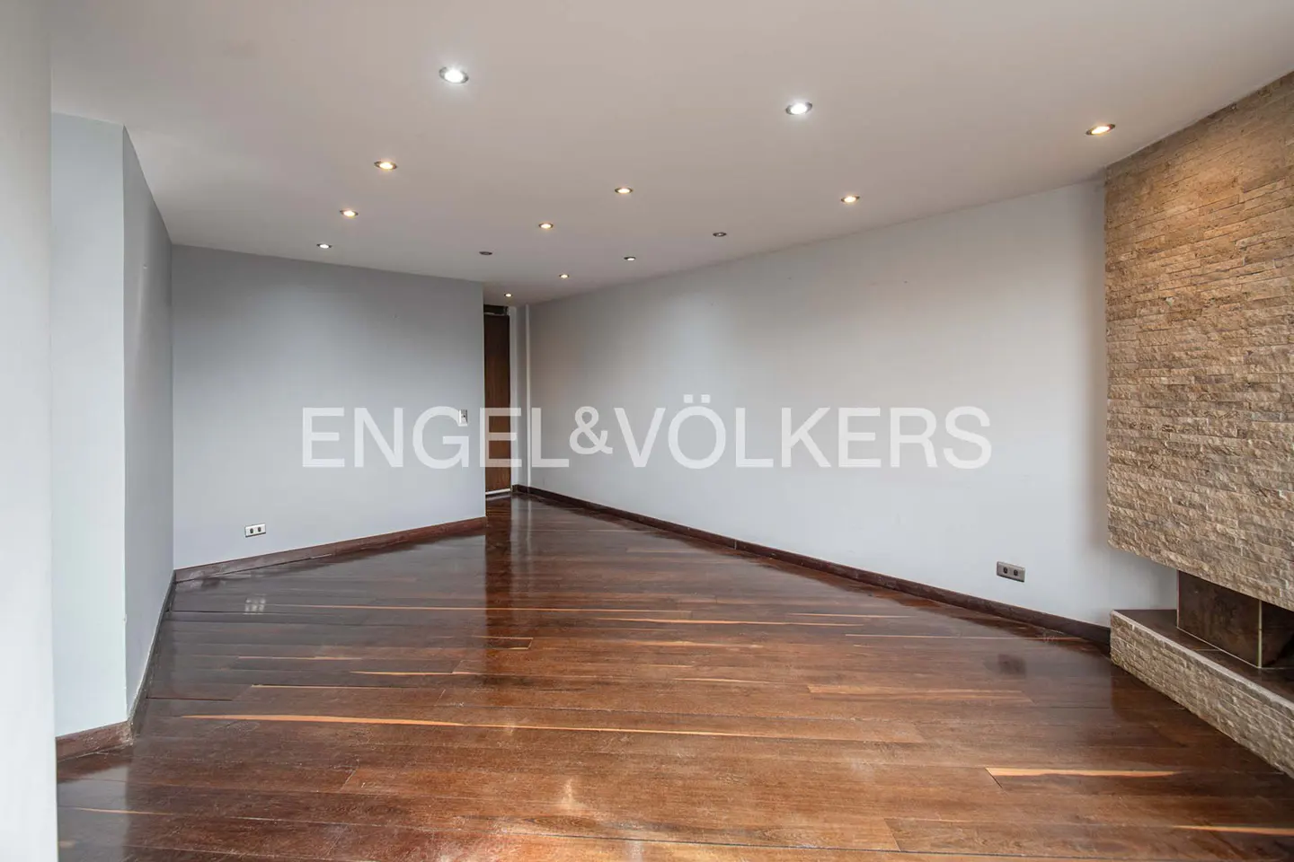 Empty living room with hardwood floors, gray walls, recessed lighting, and a stone fireplace. Engel & Völkers logo is visible.