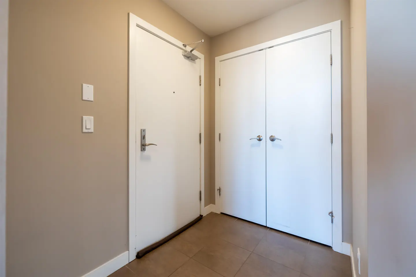 Entryway with beige walls, brown tile floor, and two white doors. One door has a silver handle and a door closer.