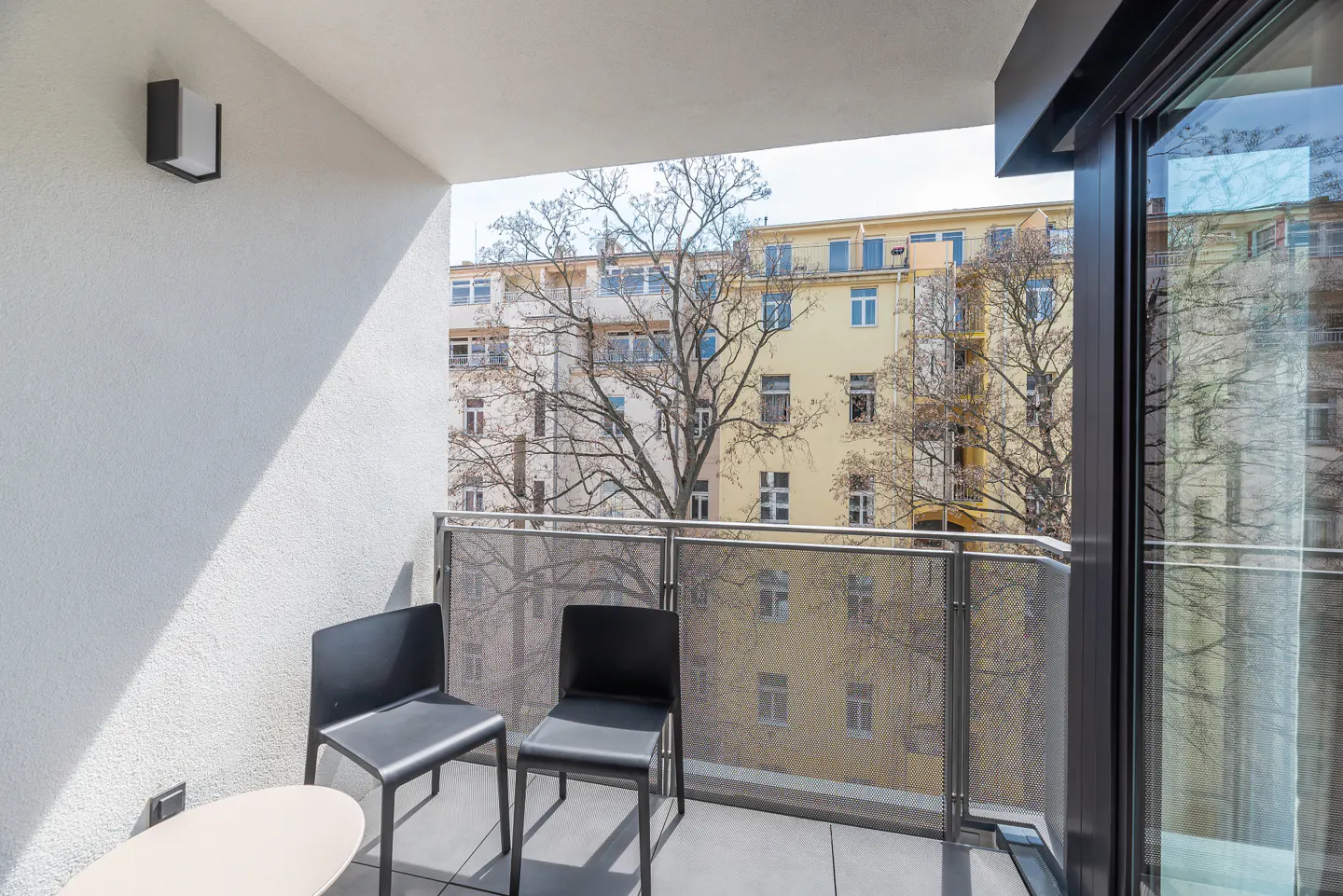 Balcony with two black chairs, a small table, and a metal railing overlooking buildings and trees.