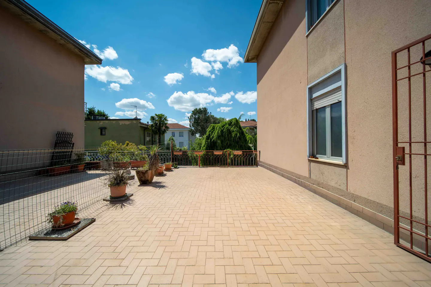 Spacious outdoor terrace with light-colored tiled flooring, potted plants, and a metal fence under a bright blue sky.