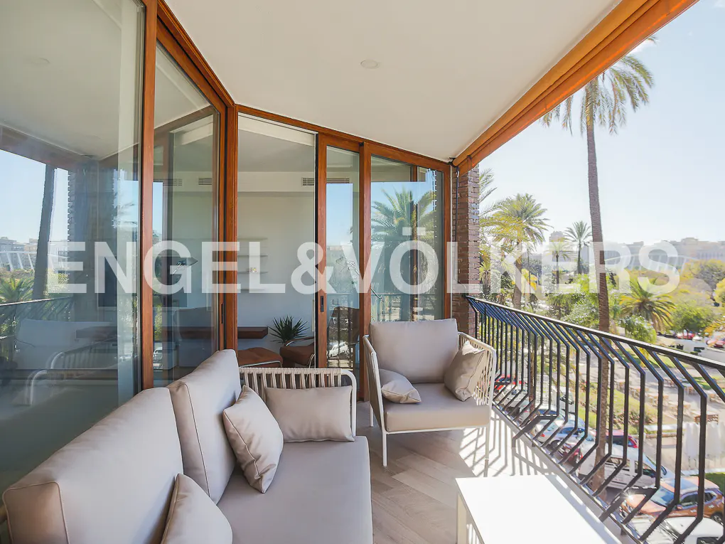 Modern balcony with beige lounge furniture, glass sliding doors, and a view of palm trees and a street below.