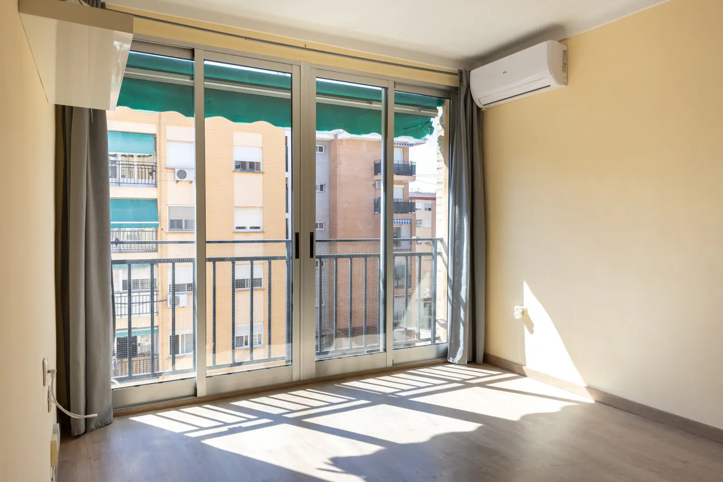 Empty room with wood floors, beige walls, and sliding glass doors to a balcony with a view of buildings. Gray curtains frame the doors.