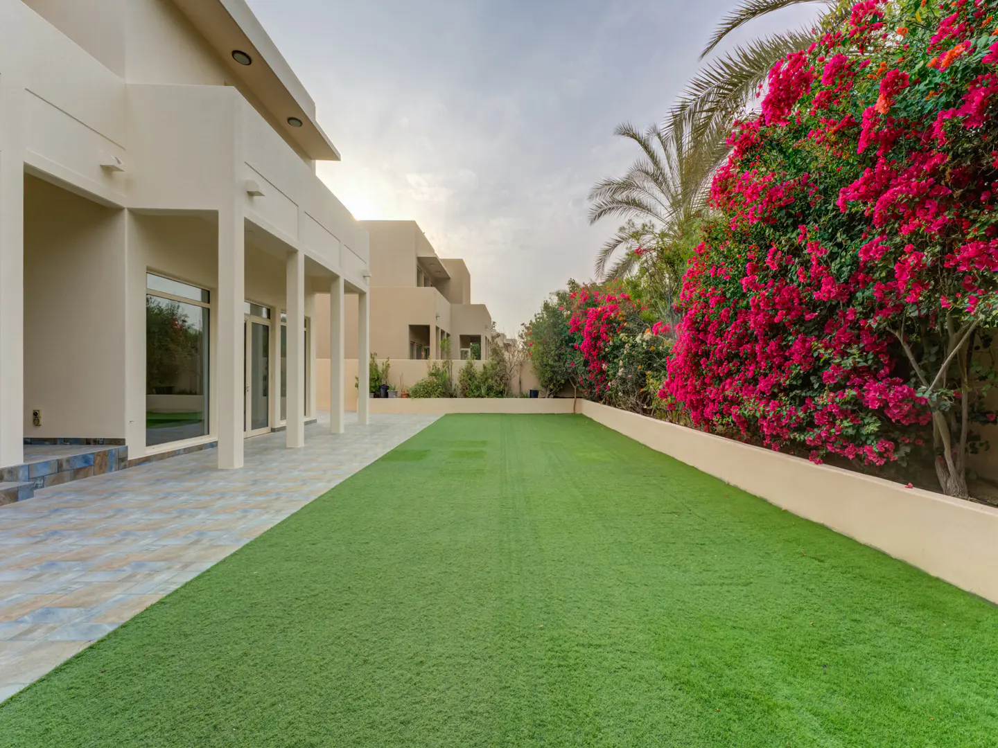 A backyard with green grass, a beige house with white pillars, and bright pink bougainvillea flowers.