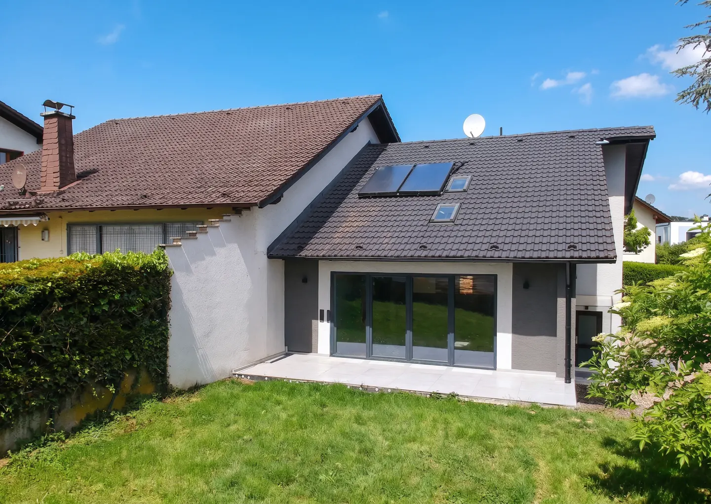 Backyard view of a house with a tiled roof, solar panels, a white patio, and large glass sliding doors under a blue sky.