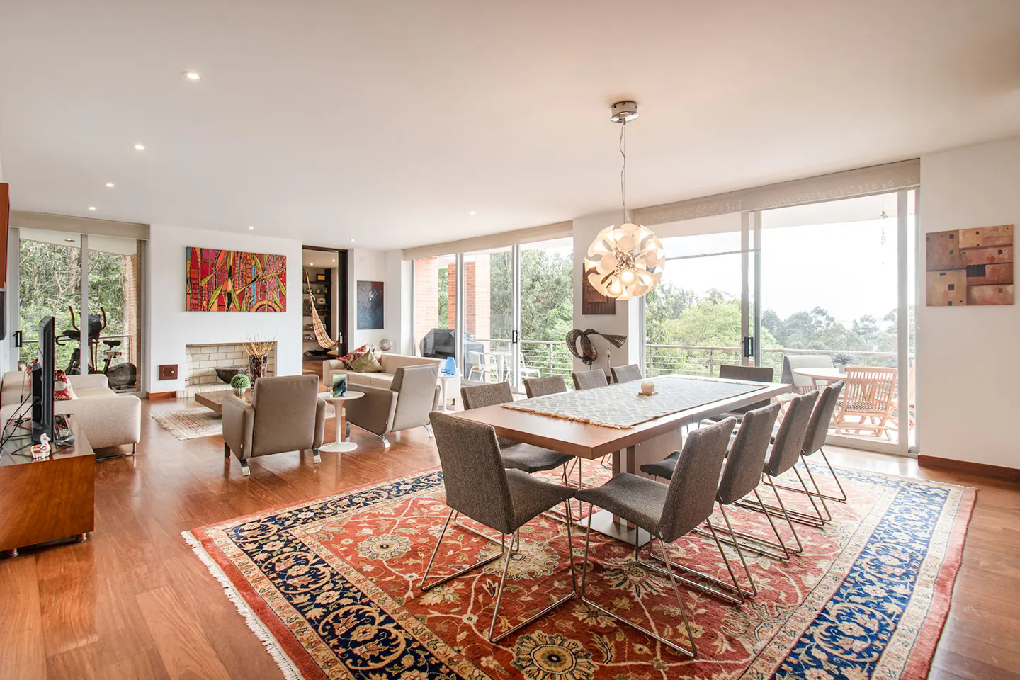 Bright, open-concept living and dining area with hardwood floors and a large red and blue patterned rug. Dining table with gray chairs, and a living room with a fireplace and colorful art. Large windows overlook greenery.