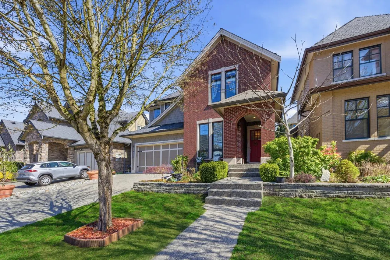 Two-story brick house with a red door, gray roof, and a stone walkway leading to the entrance. Green lawn and a tree in the front yard.