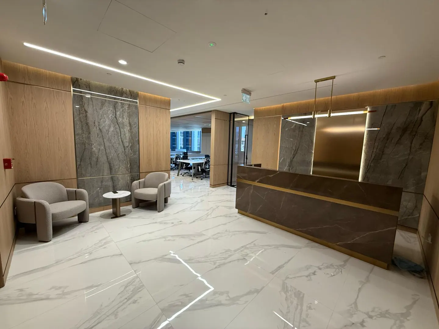 Office lobby with marble floors, wood and marble walls, and a brown reception desk. Two chairs and a small table are in the waiting area.