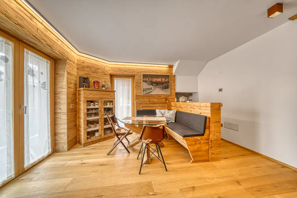 Cozy dining area with wood paneling, a glass-top table, and a built-in wooden bench with gray cushions. Two chairs are placed around the table.