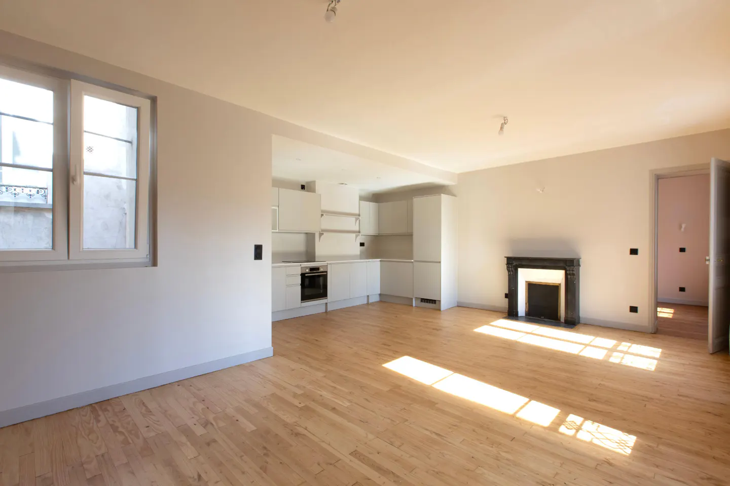 Bright, empty apartment with hardwood floors, white walls, and a black fireplace. A white kitchen is visible in the background.