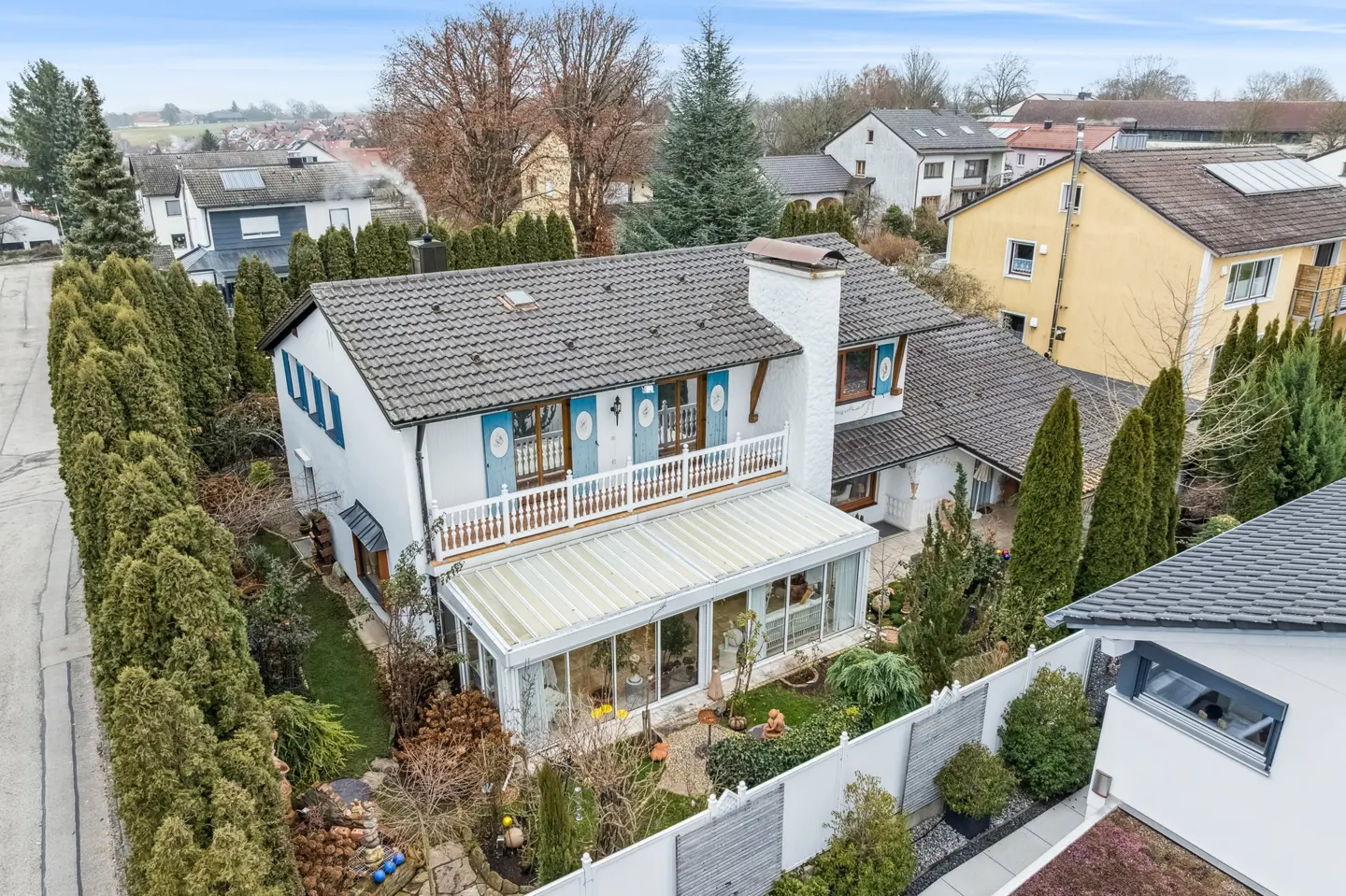 Aerial view of a white two-story house with blue shutters, a balcony, and a glass-enclosed sunroom. The house is surrounded by tall green trees and a white fence.