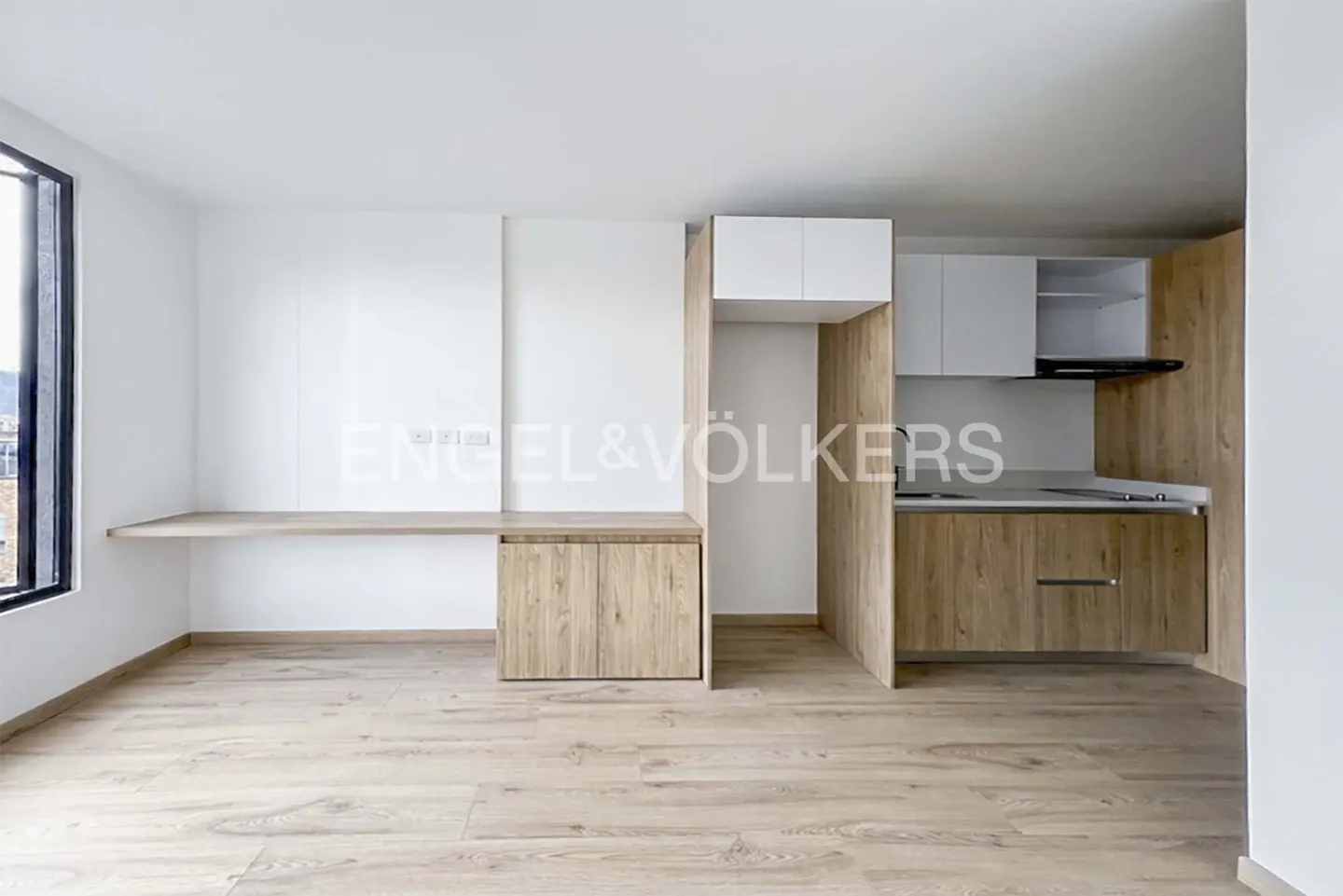 A bright, modern apartment interior with light wood floors, a built-in desk, and a kitchen with wood-grain cabinets. Engel & Völkers logo visible.