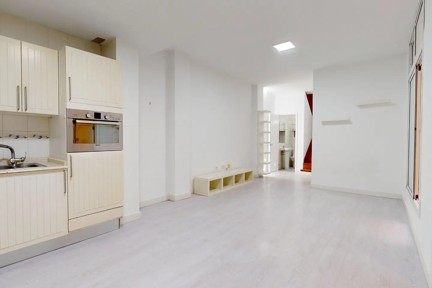 Bright, open apartment with white walls and light wood floors. Kitchen area with cabinets and stainless steel oven. Bathroom visible in the background.