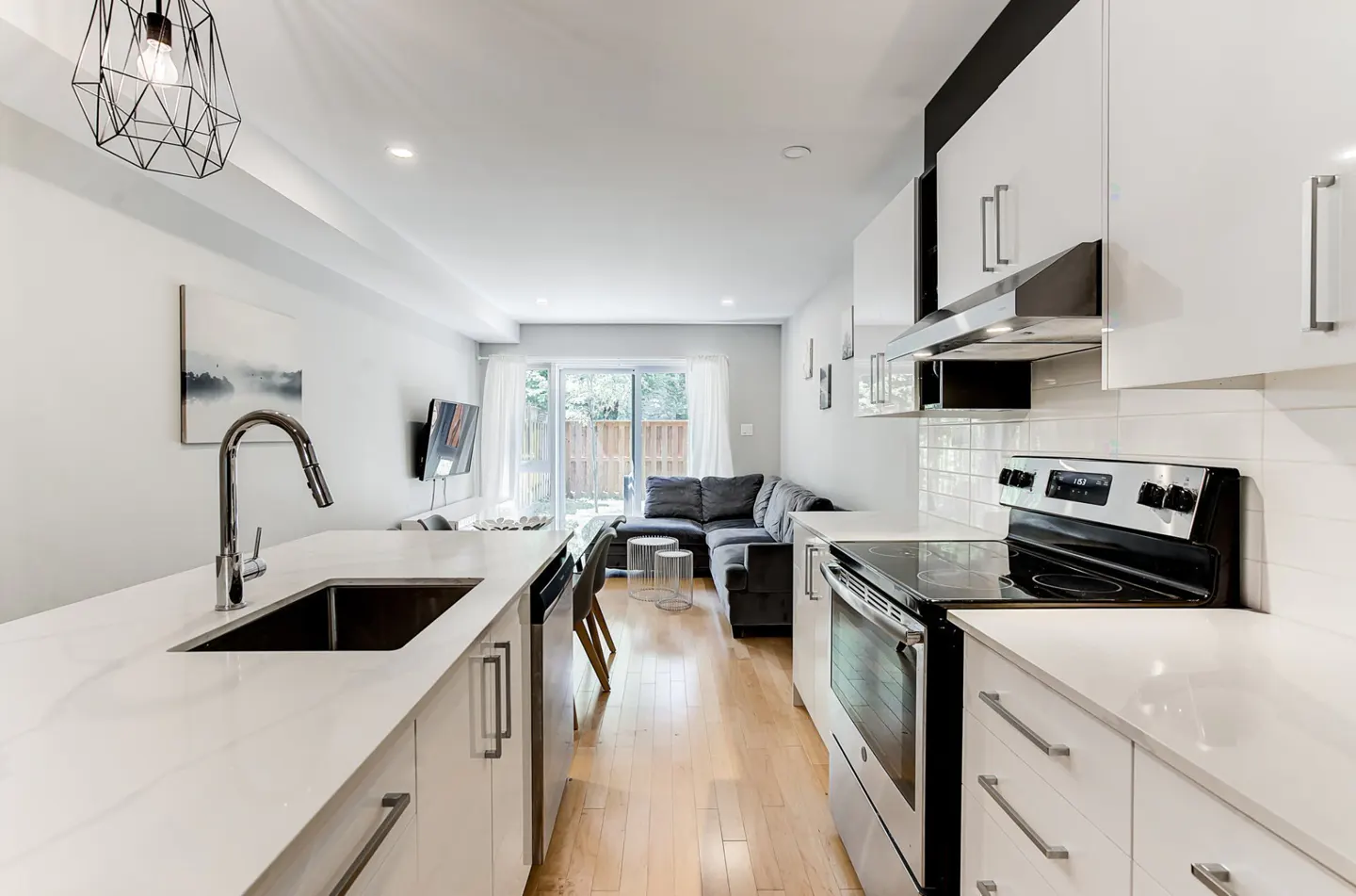 Modern open-concept kitchen with white cabinets, quartz countertops, and stainless steel appliances, leading to a living area.