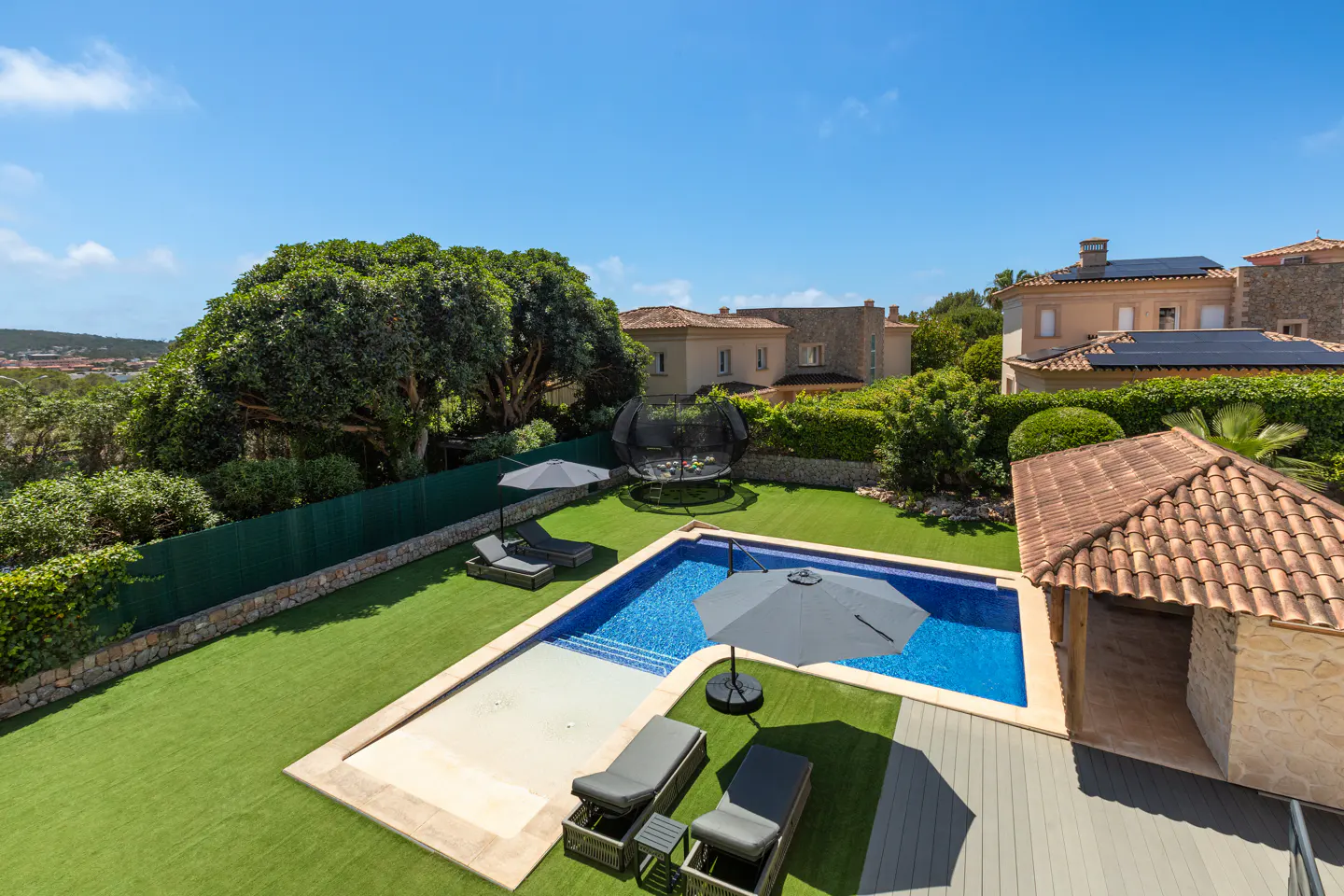Aerial view of a backyard with a blue pool, lounge chairs, umbrellas, trampoline, green lawn, and a stone pool house.