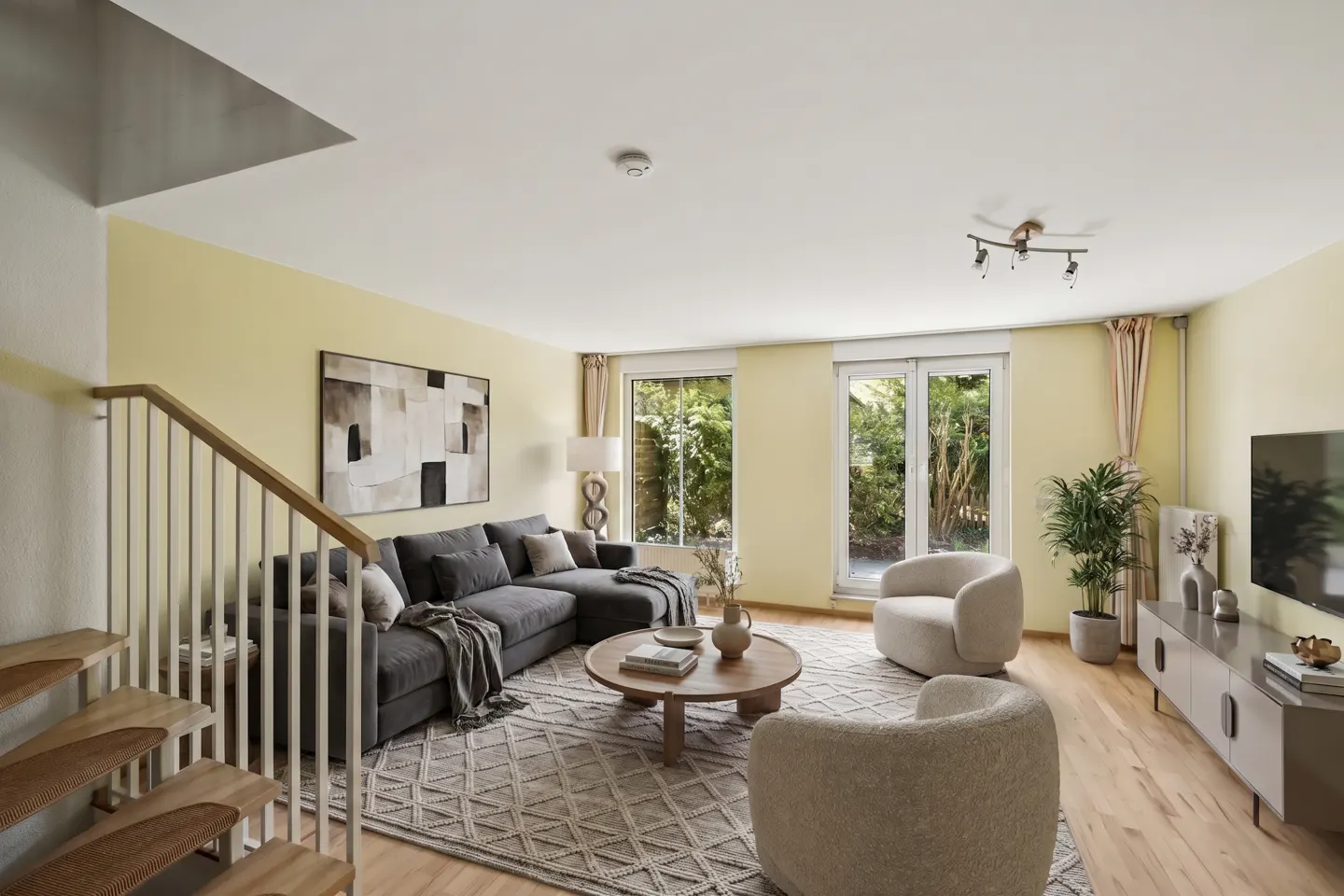 Living room with yellow walls, gray sectional sofa, two cream-colored chairs, and a wooden staircase. Windows overlook a green garden.