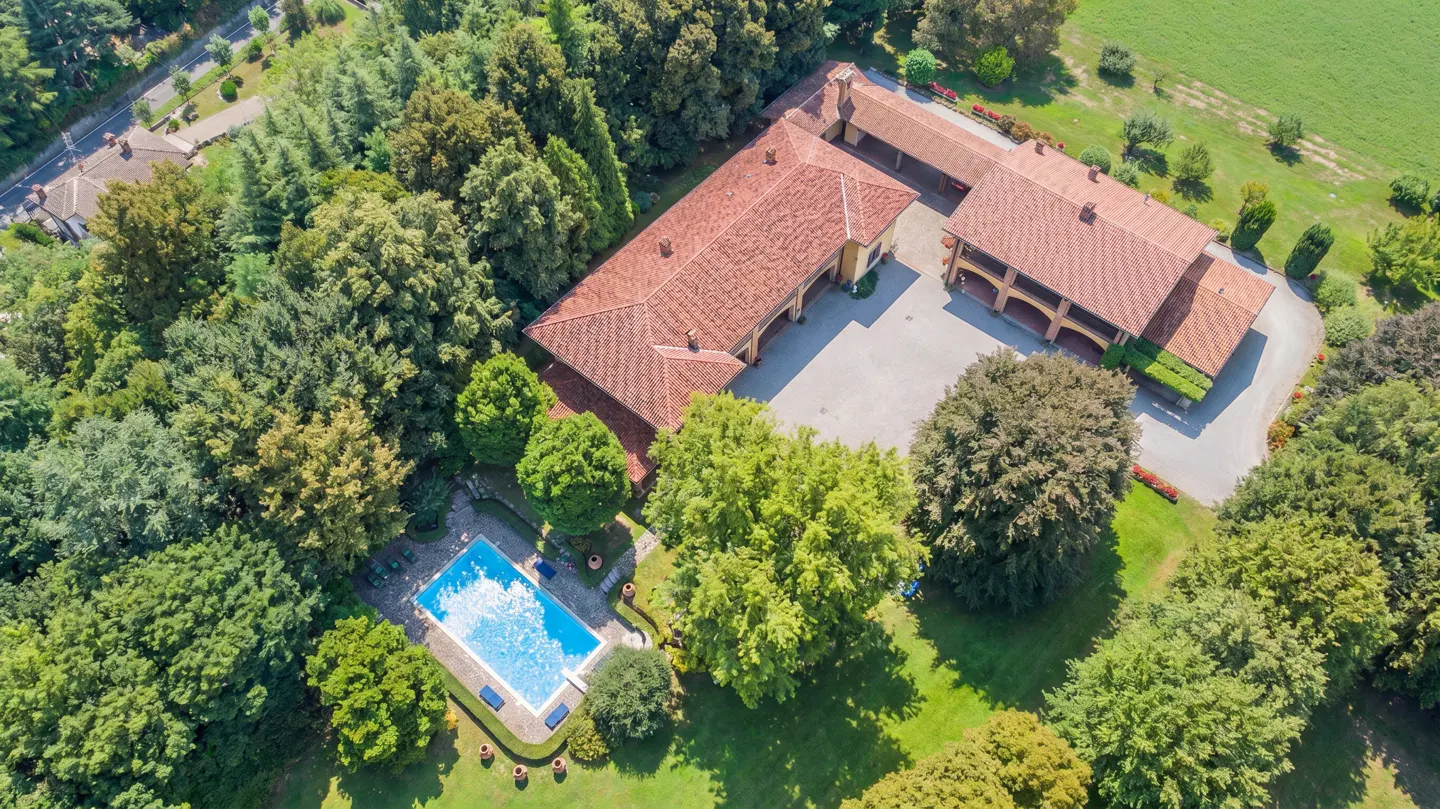 Aerial view of a large estate with a red-tiled roof, surrounded by lush green trees and a blue swimming pool.