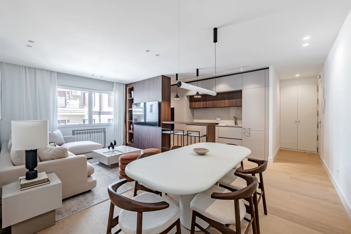 Bright, modern apartment interior with a white dining table, wood chairs, and a beige sofa. A kitchen area is visible in the background.