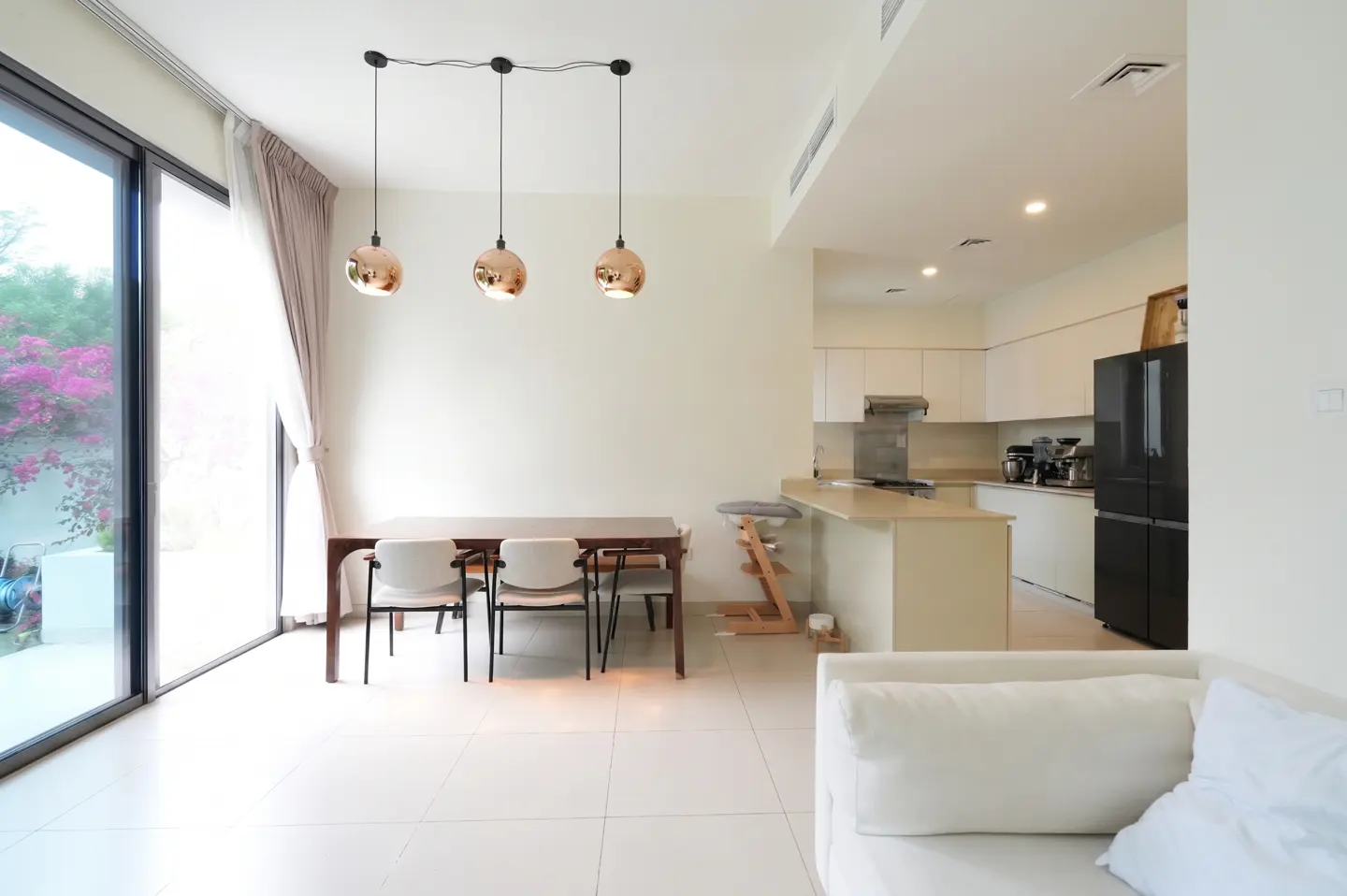 Bright, modern open-plan living space with dining table, kitchen, and white sofa. Three copper pendant lights hang above the table. Large sliding glass doors to the left.