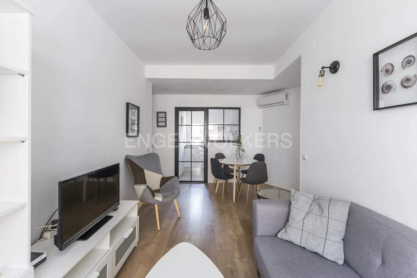 Living room with white walls, wood floors, gray sofa, TV, and dining area with black-framed glass doors.
