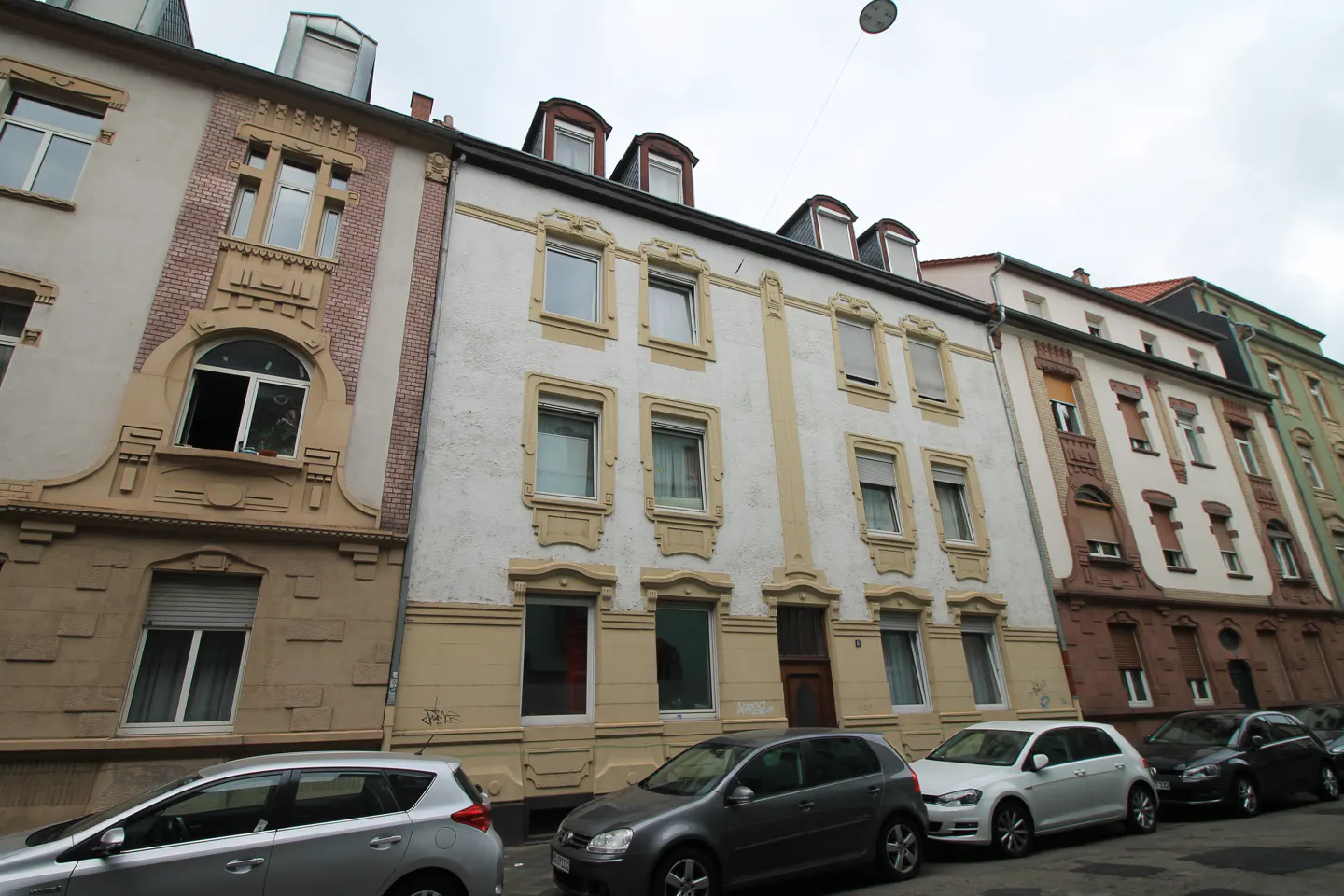 Street view of multi-story buildings with white and tan facades, dormer windows, and parked cars.