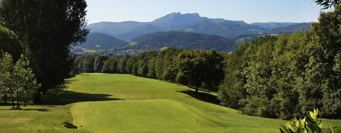 A lush green golf course fairway framed by tall trees, leading toward a scenic mountain range under a clear blue sky.