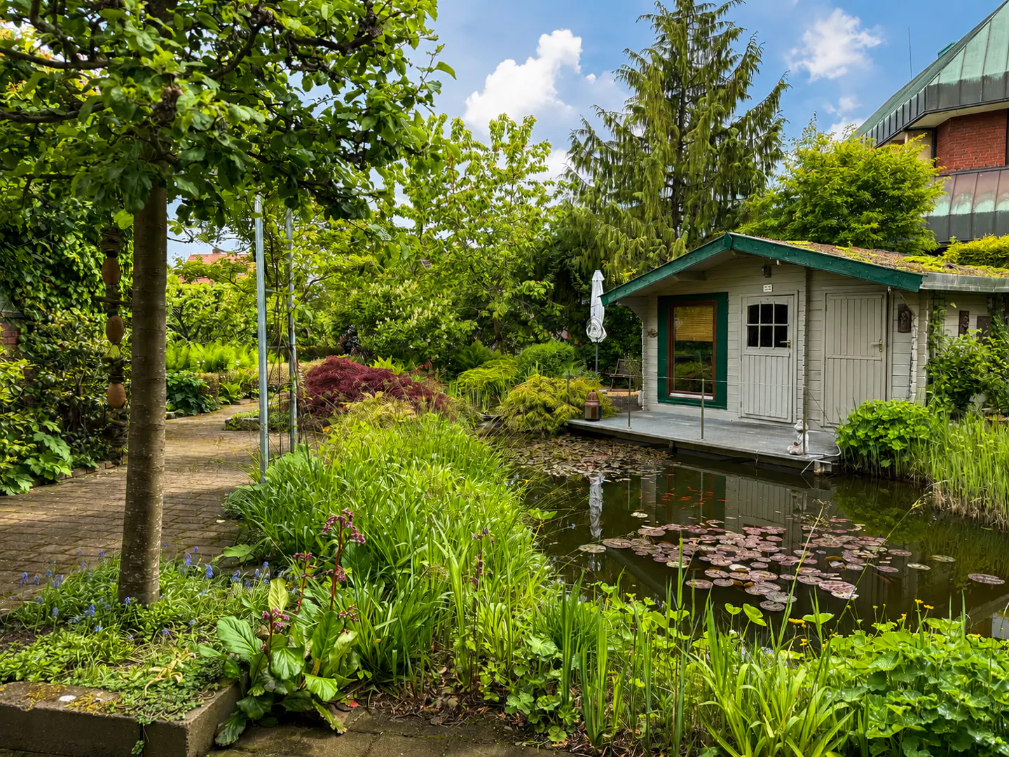 A white shed with a green roof sits on a pond with lily pads, surrounded by lush greenery and trees.