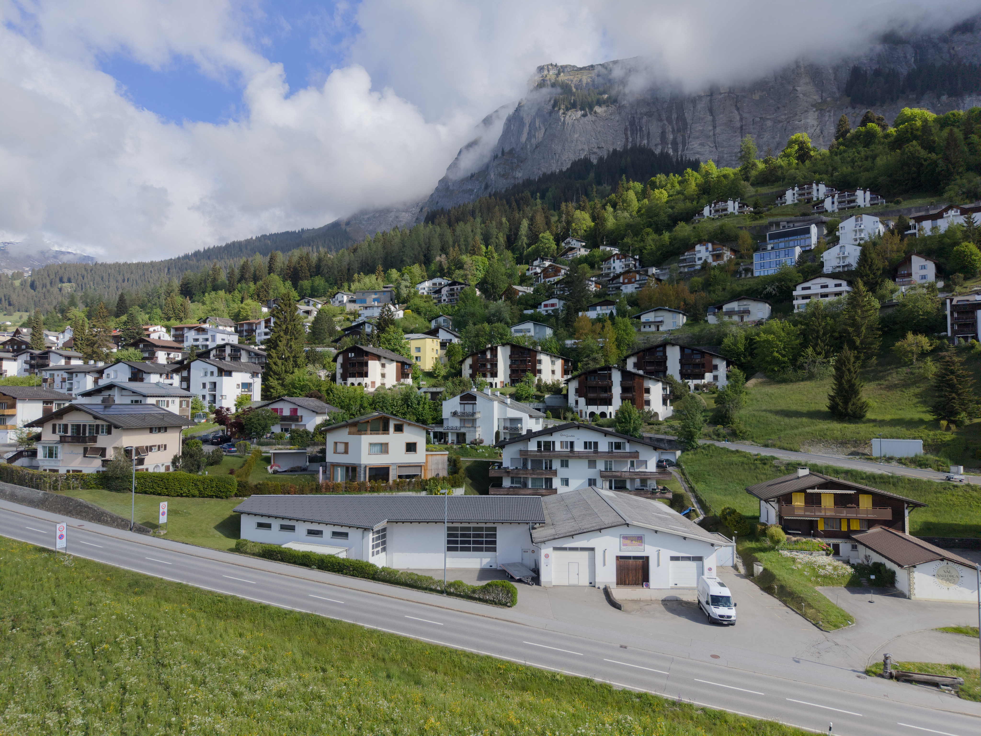 Aerial view of a Swiss village nestled on a green hillside beneath a dramatic, cloud-capped mountain peak.