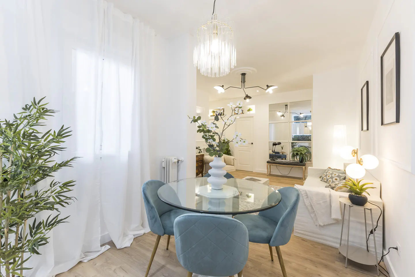 Bright, airy dining area with a glass table, four blue chairs, and a white vase with flowers. A living room is visible in the background.