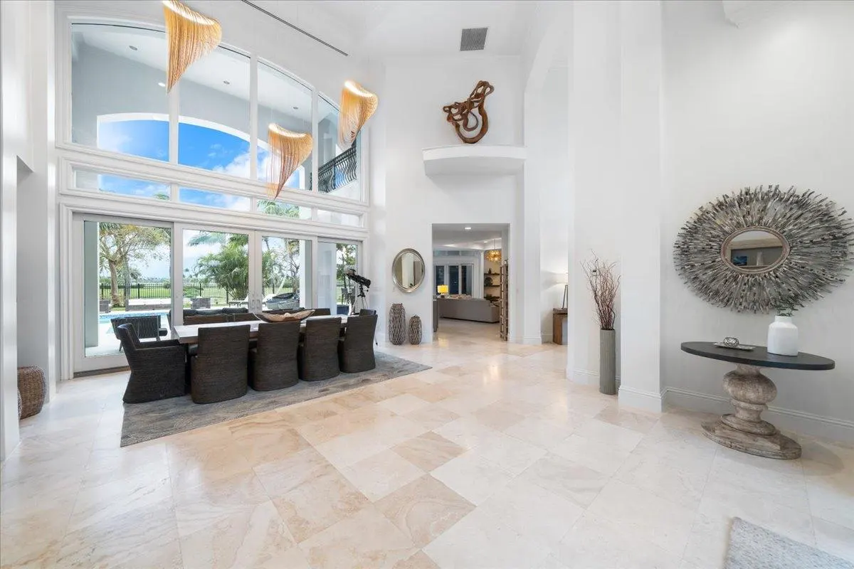 Bright, airy foyer with beige tile floors, white walls, and tall windows. A dining table sits near the windows, and a decorative mirror hangs on the wall.
