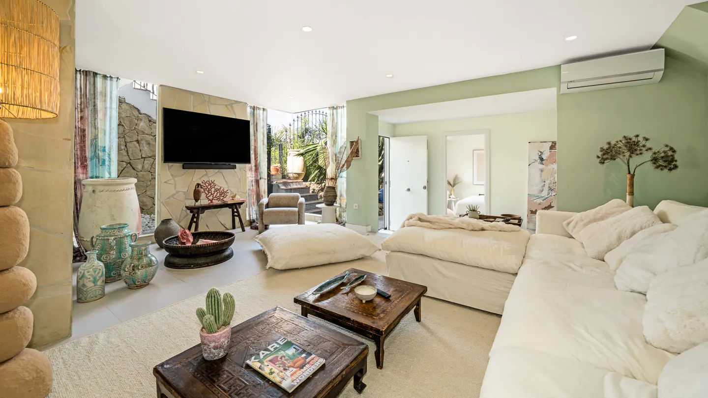 Living room with white sofa, wooden tables, and a TV. A cactus sits on a table, and a doorway leads to an outdoor area.