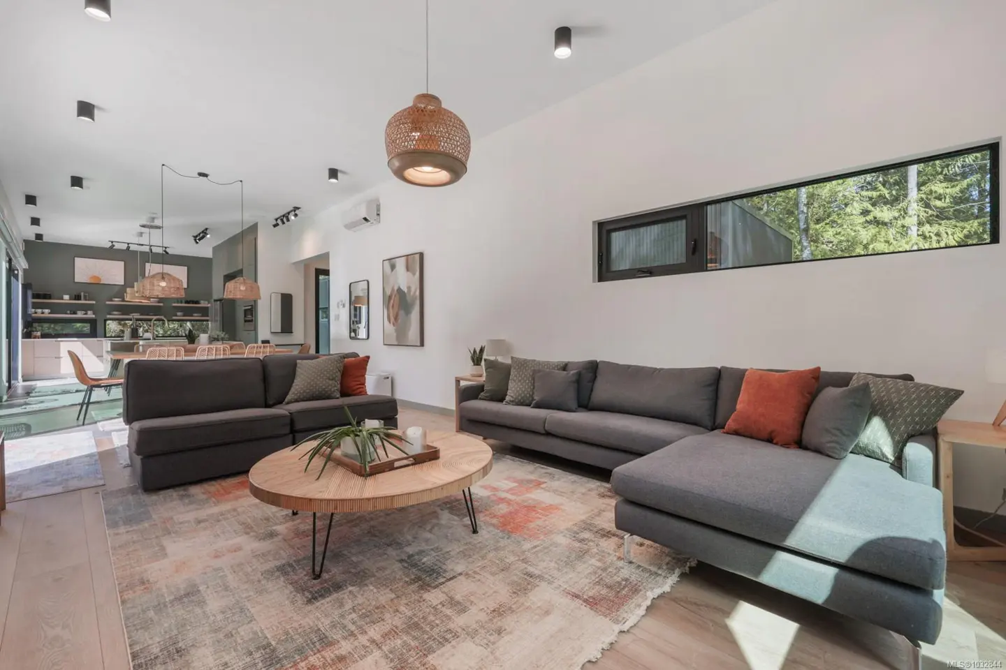 Bright, open-concept living room with gray sofas, a round wood coffee table, and a patterned rug. A kitchen and dining area are visible in the background.