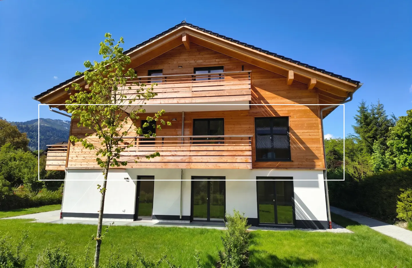 Two-story house with wood siding and white base, balconies, black windows, green lawn, and tree against a blue sky.