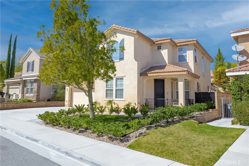 Two-story beige house with a red tile roof, green lawn, and a tree in the front yard under a blue sky.