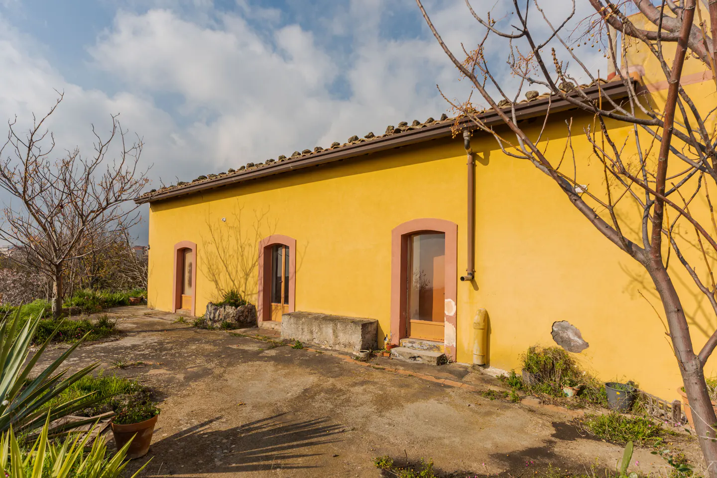 Exterior view of a yellow house with pink trim, brown roof tiles, and bare trees against a cloudy sky.